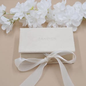 May include: A rectangular cream-colored jewelry box with a soft, textured finish. The lid features the embossed text ZANDER. A matching cream-colored satin ribbon is tied in a bow around the base of the box, which is positioned below white flowers.