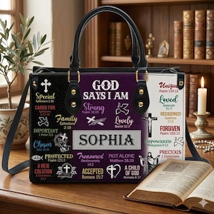Puede incluir: Bolso de mano negro, morado y blanco con asa y bandolera negras. El bolso presenta el texto "GOD SAYS I AM" y "SOPHIA", junto con palabras y símbolos religiosos. El bolso está sobre una superficie de madera.