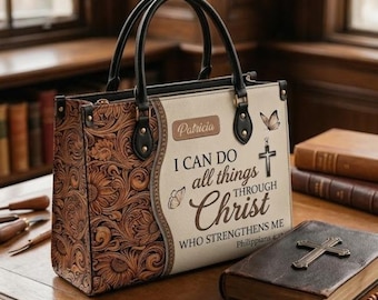 Może przedstawiać: Torba typu tote z wytłoczonym kwiatowym wzorem po bokach i napisem "I can do all things through Christ who strengthens me" z przodu. Torba ma czarne uchwyty i stoi na drewnianym stole.