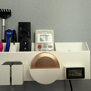 Op de afbeelding: Een witte wandorganizer met compartimenten voor diverse items. Het bevat een afstandsbediening, een klein digitaal display met temperatuur en vochtigheid, en een koperkleurige schijf. Andere items zijn pennen en een krabber.