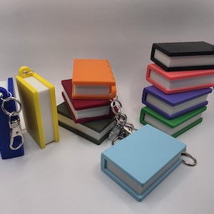 May include: A collection of miniature book-shaped keychains in various colors including blue, yellow, orange, red, green, black, pink, purple, and light blue. Each keychain features a solid-colored cover and white pages, with a metal ring attached.