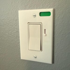 May include: A white light switch with a rectangular toggle and a small green indicator light. The switch is mounted on a gray wall and secured with two screws. A small red indicator light is also visible.
