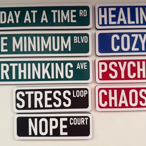 May include: A collection of rectangular signs with white text on various colored backgrounds. The signs read: "ONE DAY AT A TIME RD", "HEALING WY", "BARE MINIMUM BLVD", "COZY LANE", "OVERTHINKING AVE", "PSYCHO PATH", "STRESS LOOP", "CHAOS COURT", and "NOPE COURT".