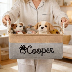 Puede incluir: Una cesta de almacenamiento rectangular con exterior gris y beige, bordada con una huella de pata y el nombre "Cooper" en negro. La cesta contiene varios juguetes de peluche para perros y tiene asas de cuerda.