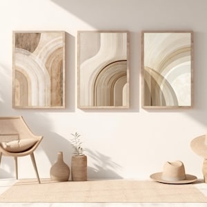 Könnte beinhalten: Drei gerahmte abstrakte Kunstdrucke in neutralen Beige- und Brauntönen hängen an einer weißen Wand. Ein Webstuhl, eine dekorative Vase und zwei Hüte befinden sich vor der Wand. Die Kunst zeigt bogenförmige Formen.