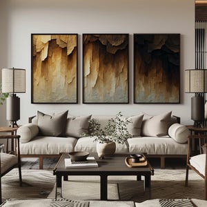 Könnte beinhalten: Ein Set aus drei rechteckigen Wandbildern mit abstrakten, geschichteten Texturen in Gold-, Braun- und Schwarztönen, gerahmt in dünnen schwarzen Rahmen über einem hellen Sofa mit neutralen Kissen und einem Couchtisch aus dunklem Holz.