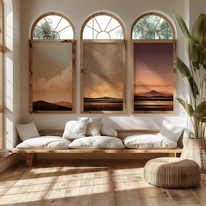 Könnte beinhalten: Drei gerahmte Landschaftskunst-Drucke mit warmen Farbpaletten hängen an einer weißen Wand unter gewölbten Fenstern. Unter den Kunstwerken befinden sich eine Holzbank mit weißen Kissen und ein gewebter Ottoman. Rechts steht eine große Topfpflanze.