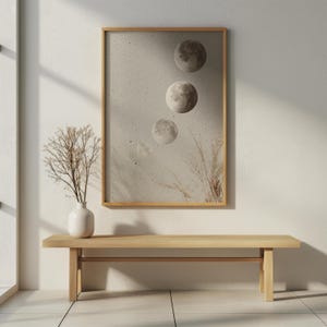 Könnte beinhalten: Ein gerahmter Kunstdruck mit drei Monden in verschiedenen Phasen vor einem strukturierten, neutralen Hintergrund. Das Kunstwerk befindet sich in einem hellen Holzrahmen, ergänzt durch eine Vase mit getrockneten Zweigen und eine Holzbank.