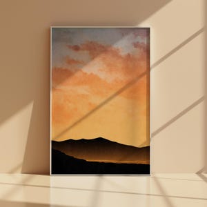 Könnte beinhalten: Ein gerahmter Landschaftskunst-Druck mit einem Sonnenuntergang über Bergen. Der Himmel wechselt von Blau zu Orange und Gelb, mit dunklen Bergsilhouetten im Vordergrund. Das Kunstwerk wird an einer neutralfarbenen Wand ausgestellt.