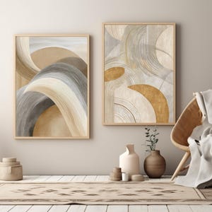 Könnte beinhalten: Zwei gerahmte abstrakte Kunstdrucke in neutralen Tönen von Beige, Grau und Gold. Das Kunstwerk zeigt fließende, geschwungene Linien und Formen. Der Raum umfasst auch einen gewebten Stuhl, einen Teppich und dekorative Vasen, die eine moderne, minimalistische Ästhetik schaffen.