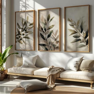 Könnte beinhalten: Drei gerahmte botanische Drucke mit Olivenzweigen und Blättern in Grün- und Brauntönen. Die Drucke werden über einem hellen Sofa mit Kissen und einer Decke präsentiert. Der Raum hat eine neutrale Farbpalette und natürliches Licht.