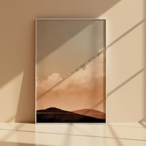 Könnte beinhalten: Gerahmter Kunstdruck mit einer minimalistischen Berglandschaft unter einem bewölkten Himmel. Das Kunstwerk verwendet eine warme Farbpalette aus Braun- und Beigetönen und erzeugt eine ruhige Ästhetik. Der Druck wird in einem weißen Rahmen präsentiert.