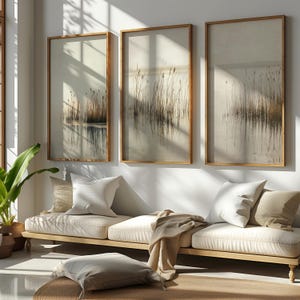 Könnte beinhalten: Drei gerahmte Landschaftskunst-Drucke, die sich im Wasser spiegelnde Schilf darstellen, in neutralen Tönen. Das Kunstwerk wird über einem hellen Holzsofa mit cremefarbenen Kissen und Kissen ausgestellt. Eine neutralfarbene Decke und eine Topfpflanze vervollständigen die Szene.