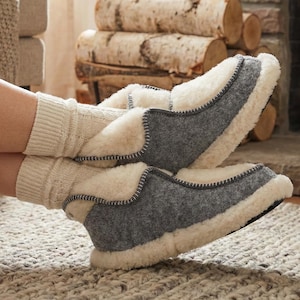 Puede incluir: Zapatillas de lana grises y crema con forro de piel de oveja. Las zapatillas tienen un exterior gris con un interior y ribete crema. La imagen sugiere comodidad y calidez.