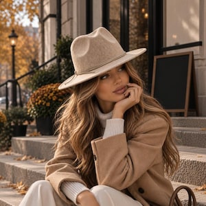 Peut inclure: Un chapeau fedora en feutre beige avec un ruban assorti, un manteau en laine marron clair, un pull à col roulé blanc côtelé et un pantalon blanc. Un sac à main structuré en cuir marron avec un fermoir doré est posé sur les marches en béton.