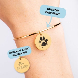 Könnte beinhalten: Goldenes Armreif-Armband mit einem Anhänger mit einem individuellen Pfotenabdruck. Ein zweiter Anhänger mit dem Text "Max 03.08.2023" ist am Armband befestigt. Der Text "CUSTOM PAW PRINT" und "OPTIONAL BACK ENGRAVING" ist auf dem Bild zu sehen.