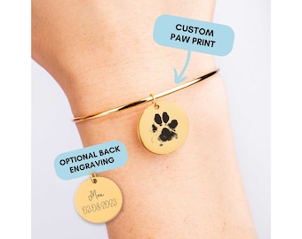 Könnte beinhalten: Goldenes Armreif-Armband mit einem Anhänger mit einem individuellen Pfotenabdruck. Ein zweiter Anhänger mit dem Text "Max 03.08.2023" ist am Armband befestigt. Der Text "CUSTOM PAW PRINT" und "OPTIONAL BACK ENGRAVING" ist auf dem Bild zu sehen.