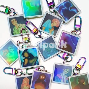 Può includere: Una collezione di portachiavi quadrati con chiusure in metallo iridescenti e colorate. Ogni portachiavi presenta un'illustrazione artistica unica in vari colori, tra cui verde, blu, rosa e viola. Le illustrazioni sembrano essere ritratti o figure.