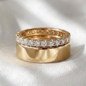 Może przedstawiać: Dwa złote pierścionki, jeden z opaską z małych, przezroczystych kamieni szlachetnych, a drugi z solidną, wypolerowaną opaską. Pierścionki są ułożone jeden na drugim, na białym, jedwabistym tle.