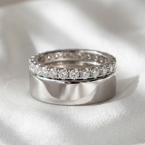 Peut inclure: Deux bagues en argent. La bague supérieure est ornée d'une rangée continue de petites pierres précieuses transparentes. La bague inférieure est une bande plus large en argent poli. Les deux bagues sont posées sur un fond blanc et soyeux.