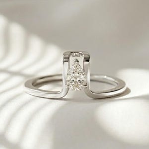 Puede incluir: Un anillo de plata con un diamante en forma de pera engastado en un marco rectangular. La banda del anillo se curva alrededor del dedo, creando un diseño moderno y elegante. El diamante brilla sobre un fondo blanco, resaltando los detalles.