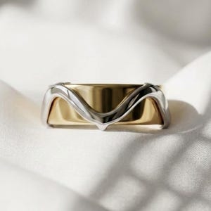 Puede incluir: Un anillo bicolor que presenta una banda lisa y pulida de color dorado, superpuesta con un elemento de metal de color plateado que forma un patrón de onda en forma de V en el centro, creando una apariencia superpuesta y con dimensión.
