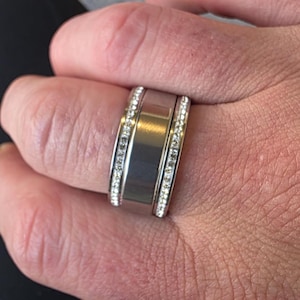 Peut inclure: Une bague argentée avec une large bande centrale plate et deux rangées de petites pierres scintillantes. La bague est portée au doigt, mettant en valeur son métal poli et l'éclat des pierres.