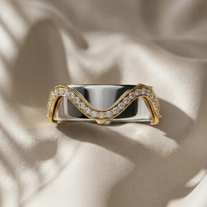 Puede incluir: Un anillo de dos tonos con una banda plateada y un diseño de onda dorada incrustado con pequeñas piedras preciosas transparentes. El anillo se encuentra sobre un fondo suave de tonos neutros, resaltando su elegante diseño. Los detalles dorados añaden un toque de lujo.