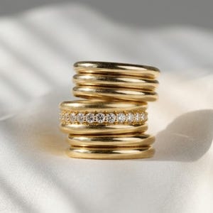 Pode incluir: Uma pilha de sete anéis de ouro dispostos verticalmente. Os anéis têm um acabamento liso e polido, e o anel central apresenta uma fileira de pequenas pedras claras de corte redondo incrustadas ao longo da sua circunferência externa.