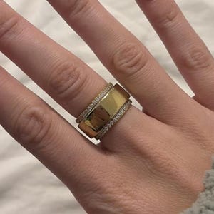 May include: A gold-colored ring with a wide band and a row of small, clear stones. The ring is on a hand with long, oval-shaped nails painted with a white tip. The background is a soft, neutral color.