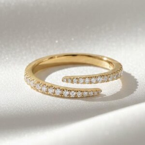 Könnte beinhalten: Ein goldfarbener Ring mit geteiltem Design, besetzt mit kleinen, klaren Edelsteinen. Der Ring ist vor einem weißen, seidigen Hintergrund platziert, der die Details des Schmucks hervorhebt. Das offene Design des Rings verleiht ihm einen modernen Touch.