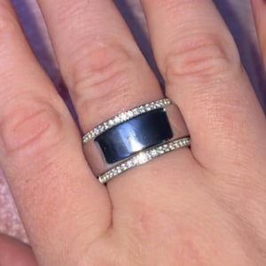 May include: A hand with pink, almond-shaped nails with silver trim. A wide, silver ring with a dark center and a row of small, clear stones on each side is worn on the ring finger.