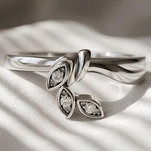 May include: A silver-toned ring featuring a bypass-style band that terminates in three leaf-shaped settings. Each leaf contains a single round, clear, faceted gemstone set within a dark, textured background, accented by small metallic beads.