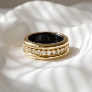 Puede incluir: Un anillo de banda ancha en oro con un canal central engastado con una hilera de piedras claras redondas de talla brillante. El interior del anillo tiene un acabado en material negro liso, creando contraste con la superficie exterior dorada.