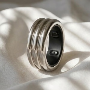 Puede incluir: Un anillo de tono plateado metálico que presenta un diseño de triple banda con una superficie exterior lisa y reflectante. El interior del anillo está forrado con un material negro mate y contiene componentes electrónicos visibles.