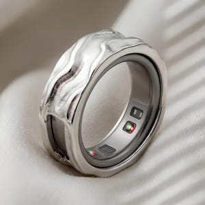 Puede incluir: Un anillo inteligente de tono plateado con una superficie exterior ondulada y texturizada y una banda interior lisa. El interior del anillo muestra sensores electrónicos integrados con pequeñas luces rojas y verdes encendidas.