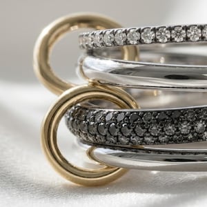 Op de afbeelding: Close-up van een verzameling ringen. De ringen zijn in goud- en zilverkleuren, sommige bezet met kleine, heldere en zwarte edelstenen. De ringen zijn in elkaar verweven, waardoor een gelaagd effect ontstaat.