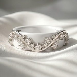 May include: A white ceramic ring featuring a silver-toned metal overlay that curves in a wave-like pattern across the front. The metal overlay is set with multiple small, round, clear faceted stones that sparkle along the entire length of the band.