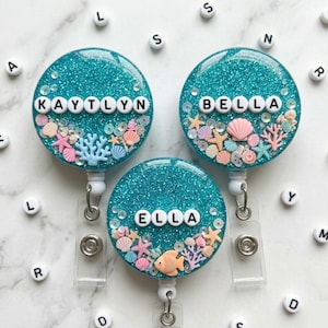 May include: Three teal glitter badge reels with the names KAYTLYN, BELLA, and ELLA spelled out in white letter beads. Each reel features miniature seashells, starfish, and coral. The reels have a clear plastic strap and a metal clip.