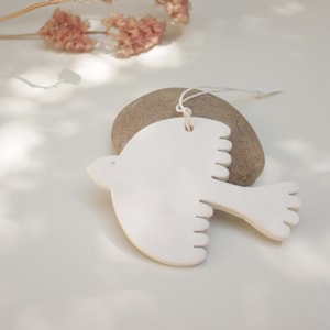 Può includere: Un ornamento bianco a forma di uccello con un cordino per appenderlo. L'ornamento è appoggiato su una pietra liscia e grigia. Fiori rosa secchi sullo sfondo e la scena è illuminata dalla luce solare soffusa.