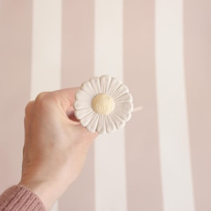 Op de afbeelding: Een madeliefvormige kastknop met een crèmekleurige buitenkant en een gele kern. De knop wordt in een hand gehouden tegen een achtergrond van verticale roze en witte strepen. Deze decoratieve hardware voegt een bloemig tintje toe aan meubels.