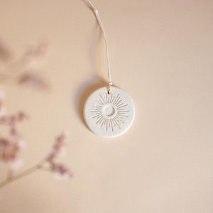 Puede incluir: Adorno redondo de cerámica blanquecina con diseño de sol. El sol presenta un círculo central con líneas radiales. Una fina cuerda blanca está unida para colgarlo. El fondo es de un color suave y neutro.