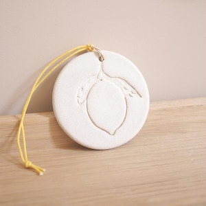 Peut inclure: Ornement rond en céramique blanche avec un motif de citron gravé. Le citron est orné de feuilles. Une ficelle jaune est attachée pour suspendre l'ornement. Il est posé sur une surface en bois.