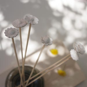 Peut inclure: Un ensemble de bâtonnets diffuseurs blancs en forme de fleurs dans un flacon en verre transparent. Les têtes de fleurs présentent des motifs de pétales détaillés. Les bâtonnets en bois sont fins et de couleur claire, créant une esthétique naturelle.