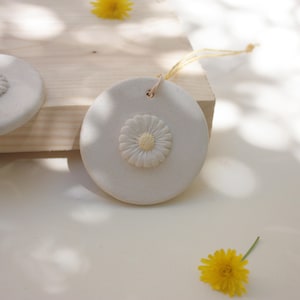 Puede incluir: Adorno redondo de cerámica blanquecina con un diseño de margarita en relieve en el centro. La margarita tiene un centro amarillo y pétalos blancos. Un pequeño agujero en la parte superior para colgar con una cuerda amarilla. Aproximadamente 7.6 cm de diámetro.