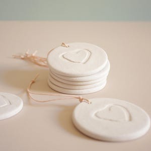 Peut inclure: Une pile de six ornements ronds en céramique blanche avec une empreinte en forme de cœur. Chaque ornement a un petit trou pour l'enfilage. Quelques ornements sont éparpillés, avec une ficelle rose attachée.