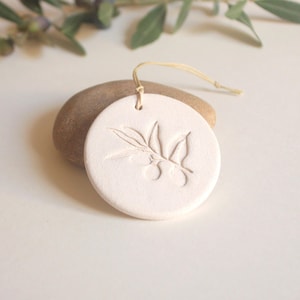 Peut inclure: Ornement rond en céramique blanc cassé avec un motif de branche d'olivier en relief. Il possède un petit trou en haut avec une ficelle beige pour l'accrocher. L'ornement repose sur une pierre lisse et brune, avec des branches d'olivier en arrière-plan.