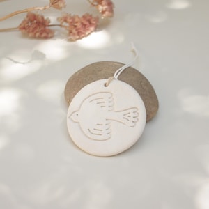 Peut inclure: Ornement rond en céramique blanc cassé avec un motif d'oiseau en vol sculpté. L'ornement est suspendu à une ficelle blanche et repose sur une pierre grise lisse. Des fleurs roses séchées sont en arrière-plan.