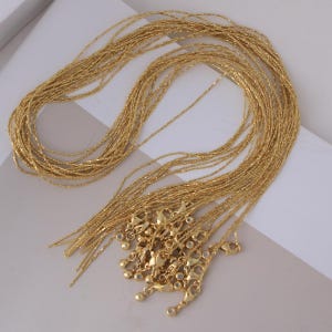 Könnte beinhalten: Eine Sammlung goldfarbener Halsketten mit zarten, strukturierten Ketten. Jede Halskette verfügt über einen Karabinerverschluss und kleine, dekorative Endperlen. Die Halsketten sind in einem lockeren, überlappenden Muster angeordnet.