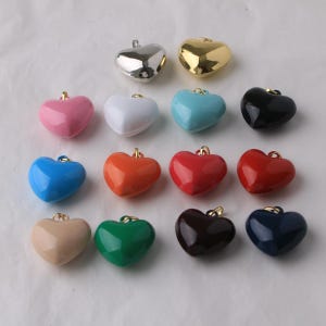 Puede incluir: Una colección de dijes en forma de corazón en varios colores, incluyendo plata, oro, rosa, blanco, azul, negro, naranja, rojo, beige, verde y azul marino. Cada dije tiene un pequeño lazo dorado en la parte superior para sujetar.